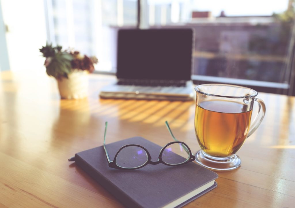 Photo of a home-office desk with notebook, glasses, cup of tea and laptop.