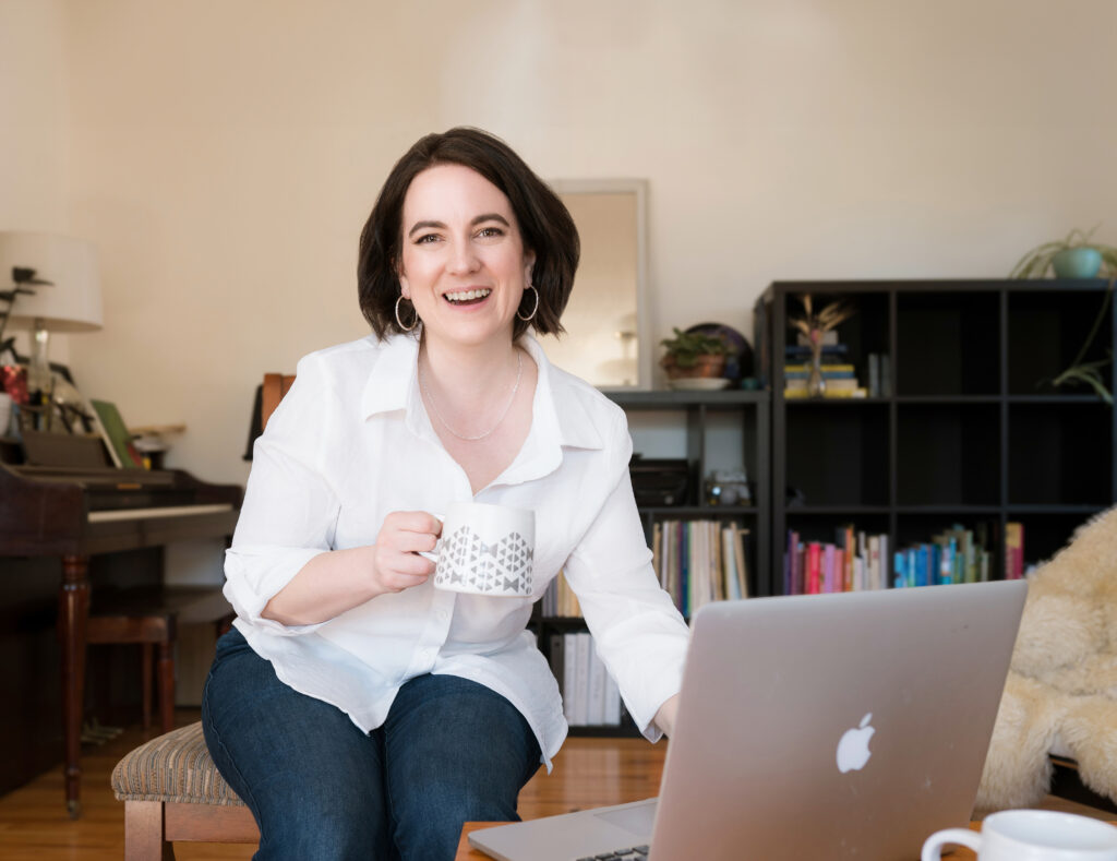 Photo of Angelica in living room. She is seated by a laptop, holding a cup up too, and giving you a big welcoming smile.