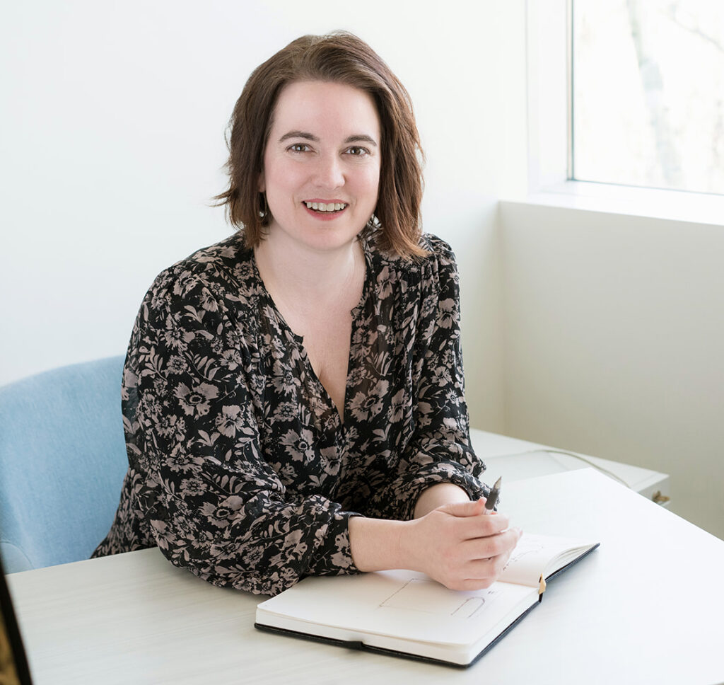 Angelica Hesse, leaning on a note book. She is looking at you with an enthusiastic smile.